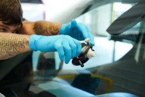 Person with tattooed arms and blue gloves repairing a chip on a car windshield using a specialized tool